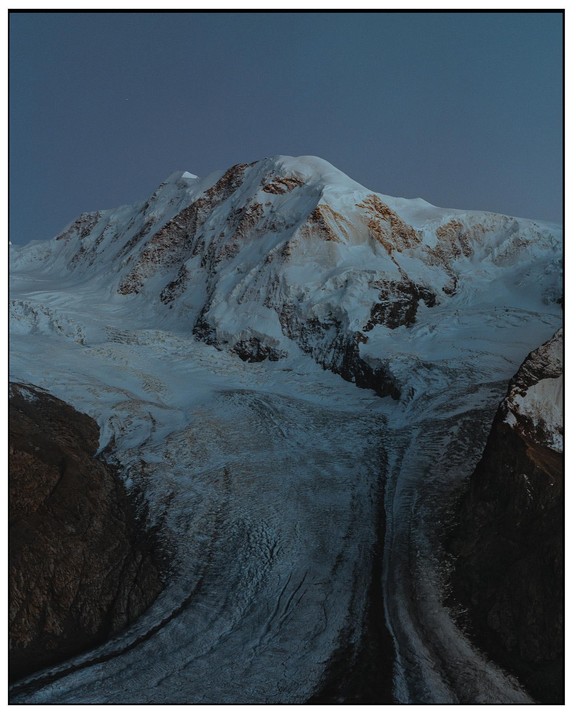A snow-covered mountain peak rises above a glacier at dusk.