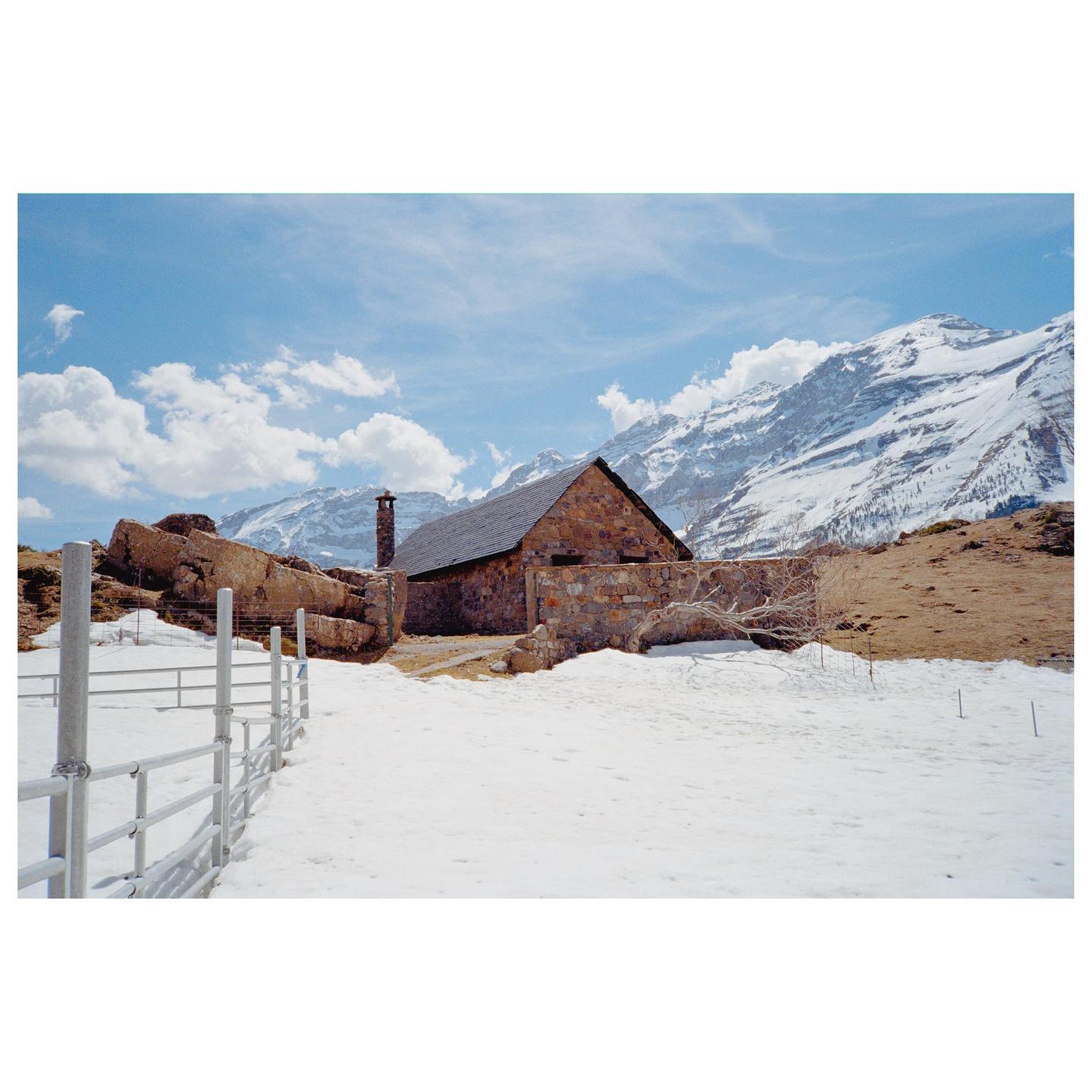 A stone building with a dark roof and chimney stands in a snowy landscape with mountains in the background.