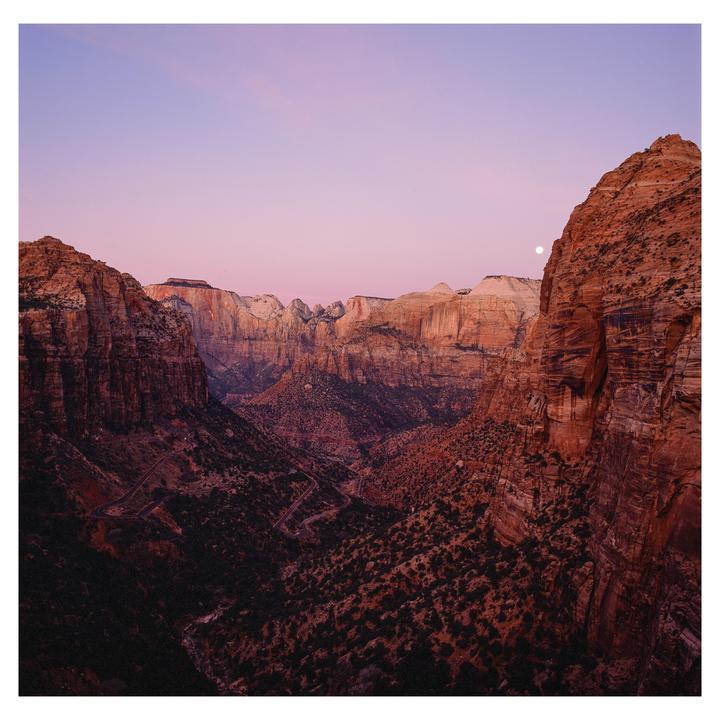A full moon is visible over a canyon landscape with a winding road at dusk.