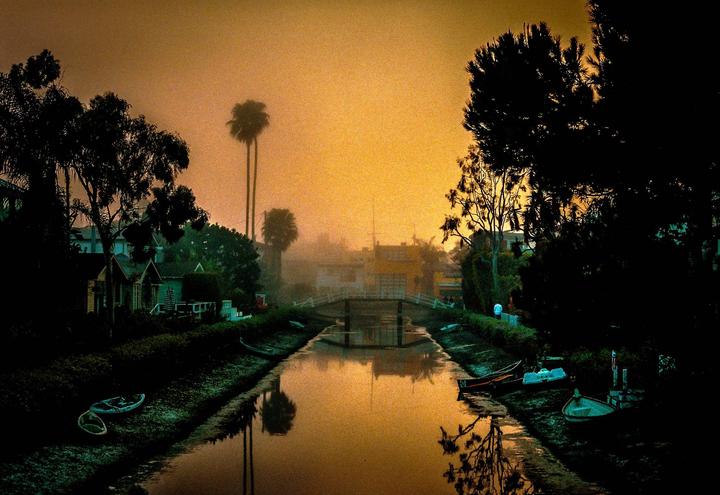 A canal with a bridge and houses on its banks is shrouded in fog at sunset.