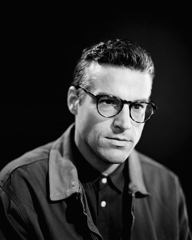 A man wearing glasses and a jacket looks directly at the viewer.