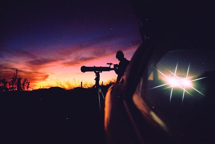 A person looks through a telescope at dusk with a car in the foreground.