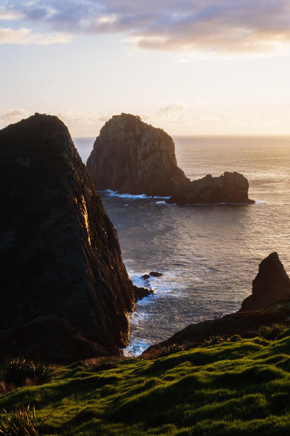 The sun rises over the ocean, illuminating a rocky coastline with a large sea stack in the distance.