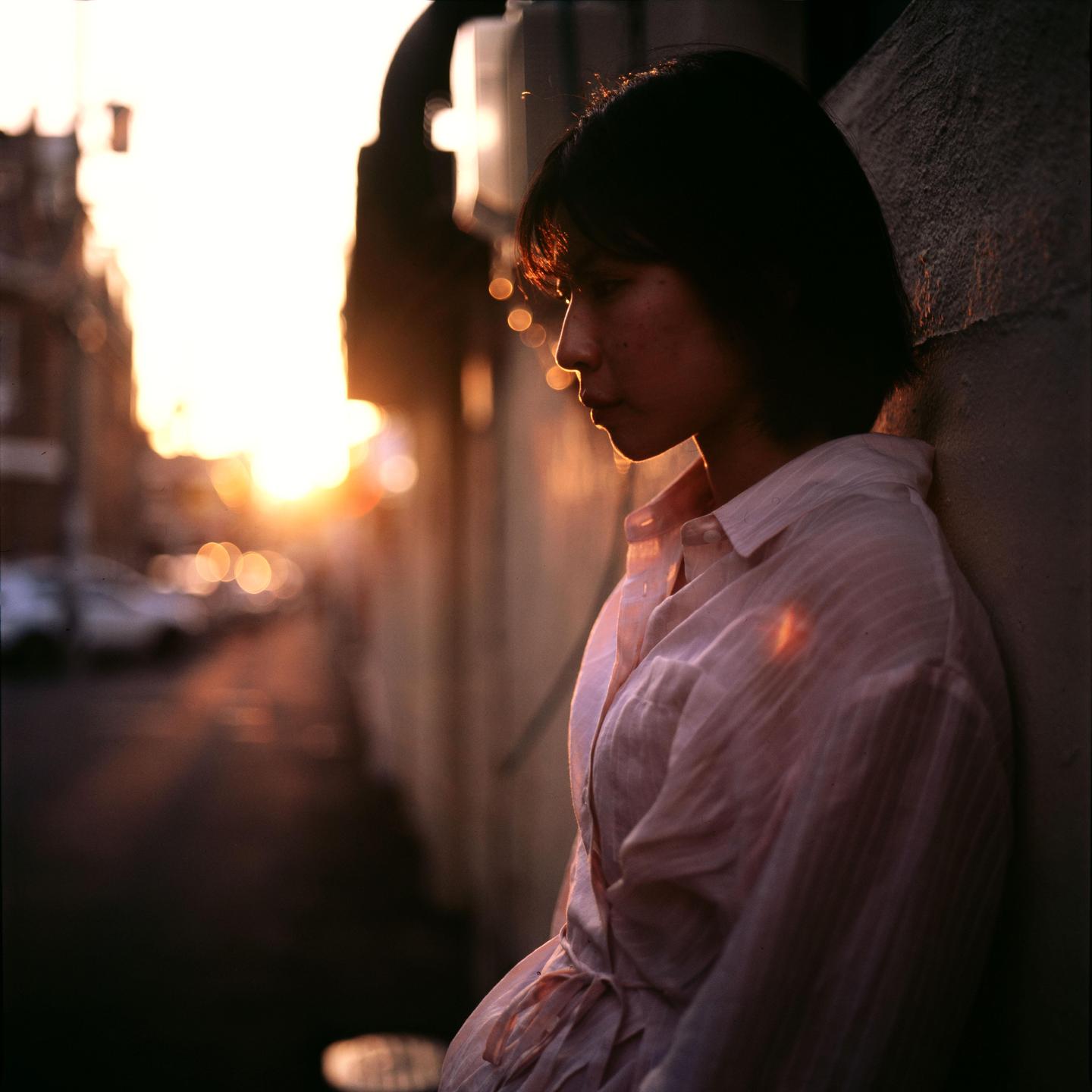 A woman stands in profile against a wall with a bright sunset in the background.