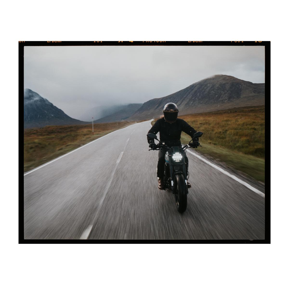 A person on a motorcycle rides down a road with mountains in the background.