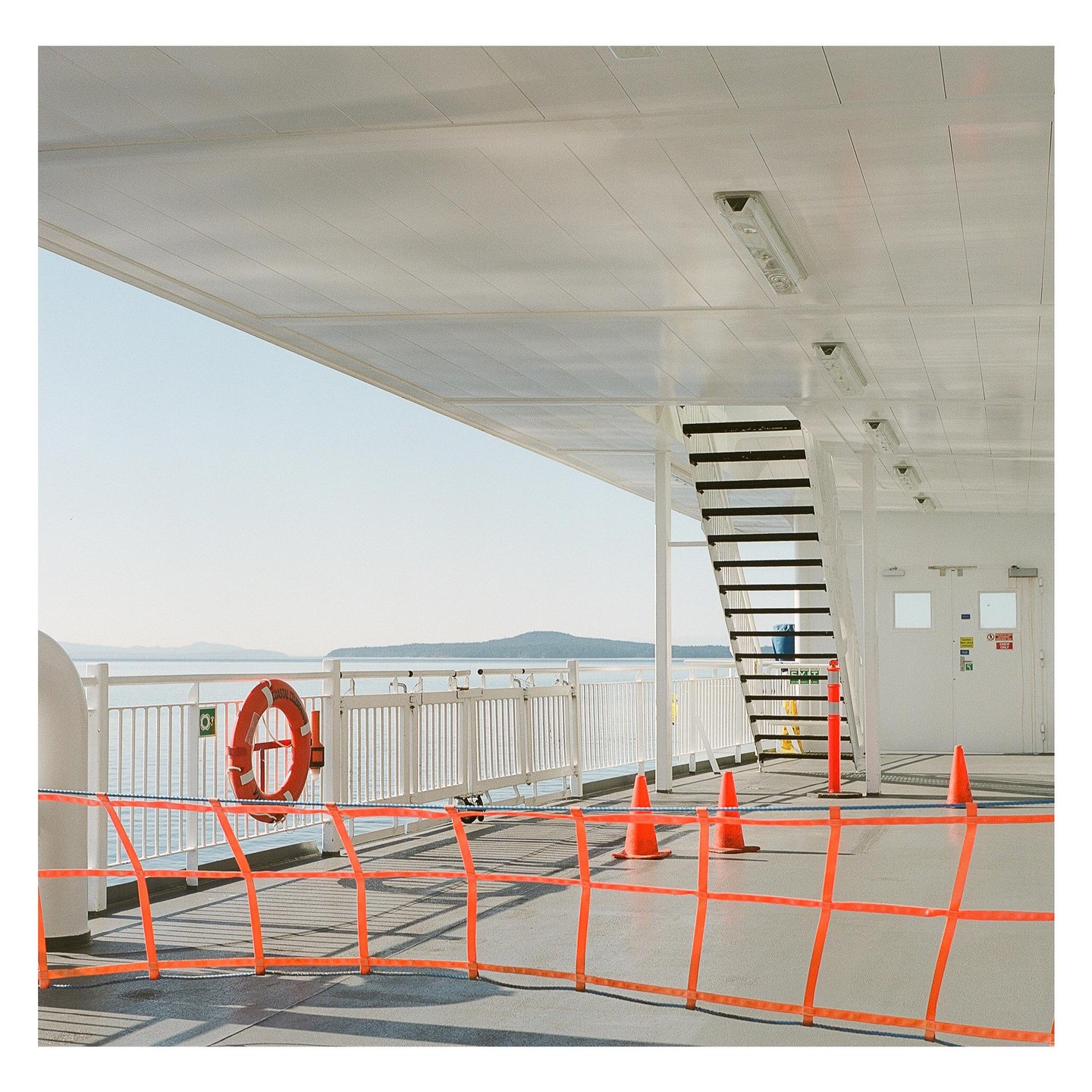 A ferry deck with orange safety barriers and a life preserver overlooks a body of water with distant islands.