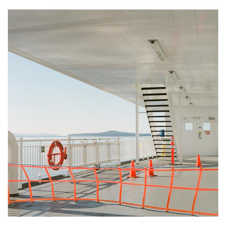 A ferry deck with orange safety barriers and a life preserver overlooks a body of water with distant islands.
