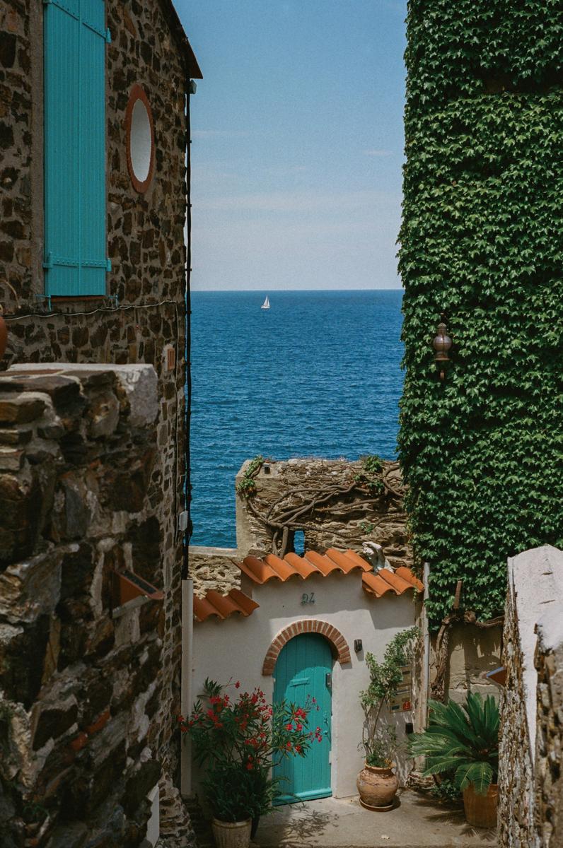 A narrow alleyway between stone buildings with a turquoise door and a view of the sea with a sailboat in the distance.