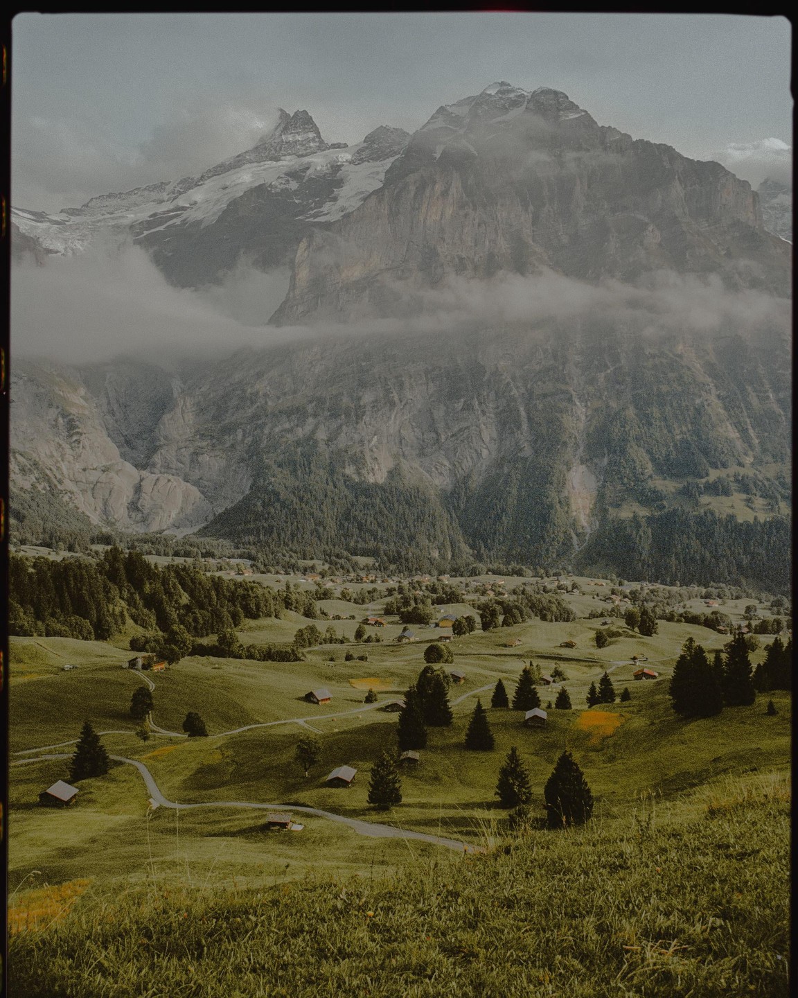A valley with small houses and trees is nestled between large, cloud-covered mountains.