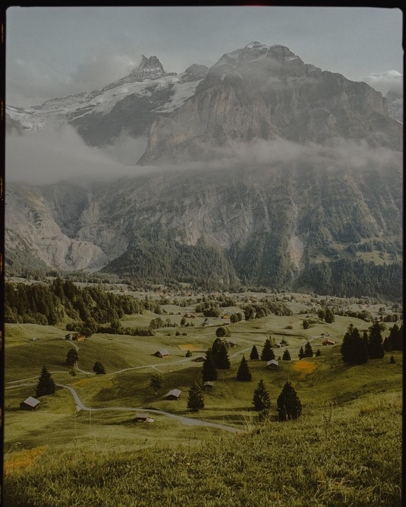 A valley with small houses and trees is nestled between large, cloud-covered mountains.