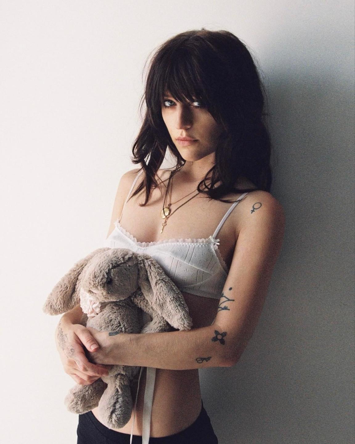 A woman with dark hair and bangs wears a white bra and holds a stuffed bunny against a white wall.