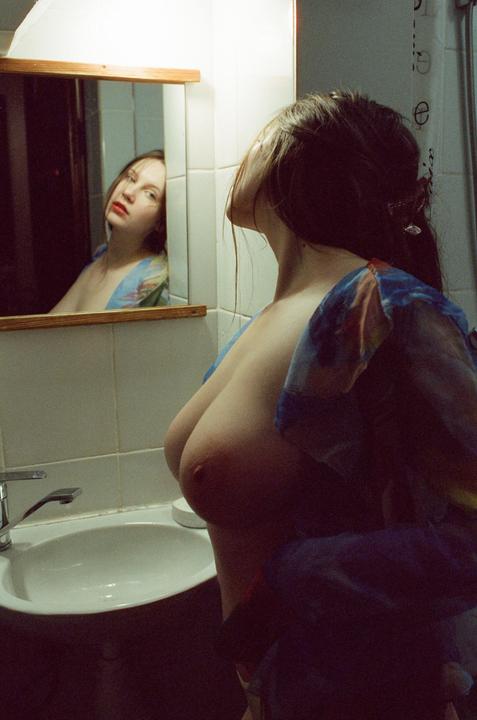 A woman stands topless in a bathroom, looking at her reflection in a mirror.