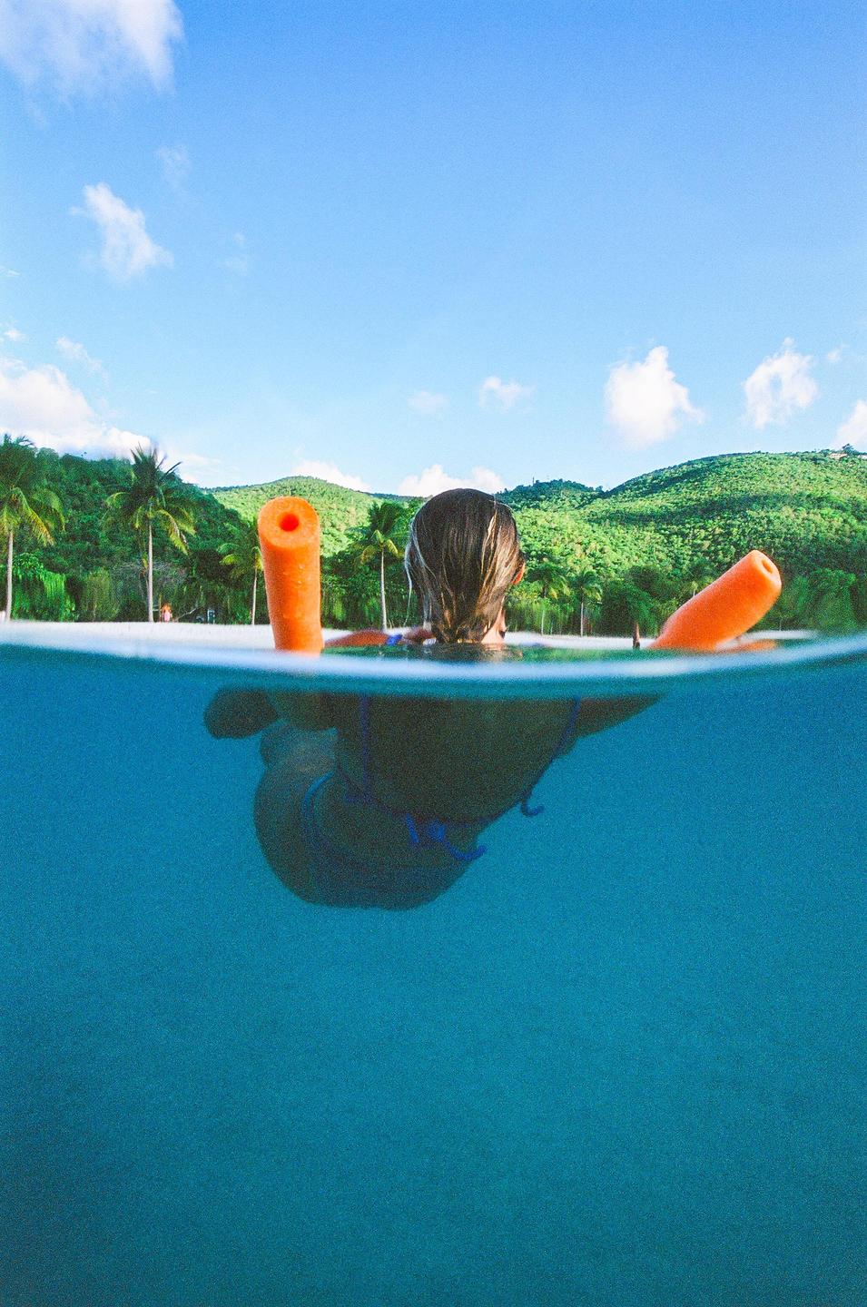 A woman floats in the ocean with two orange pool noodles, looking towards a tropical beach with palm trees and green hills.