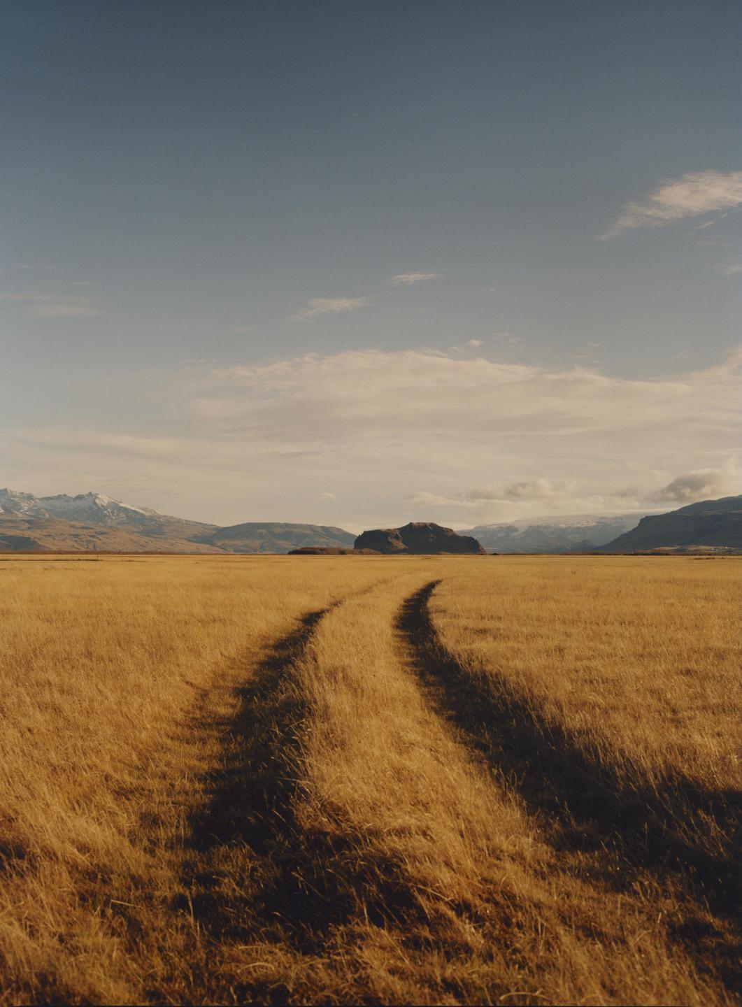 A dirt track curves through a field of golden grass towards mountains under a partly cloudy sky.