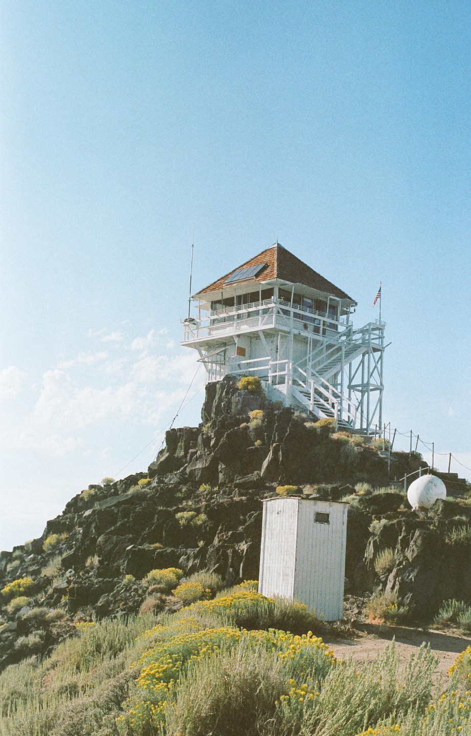 A white lookout tower with a brown roof stands on a rocky hill covered in green and yellow vegetation under a clear blue sky.