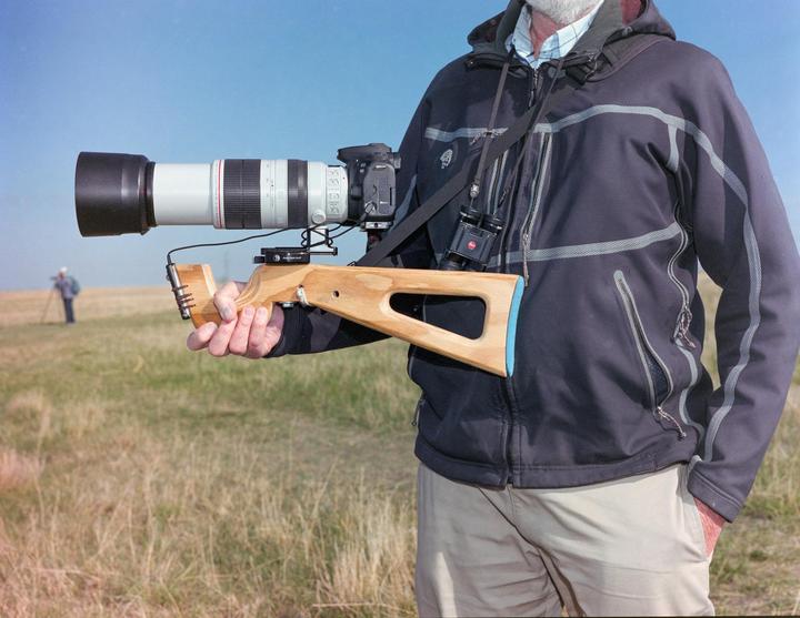 A man holds a camera with a large lens attached to a wooden stock in a grassy field.