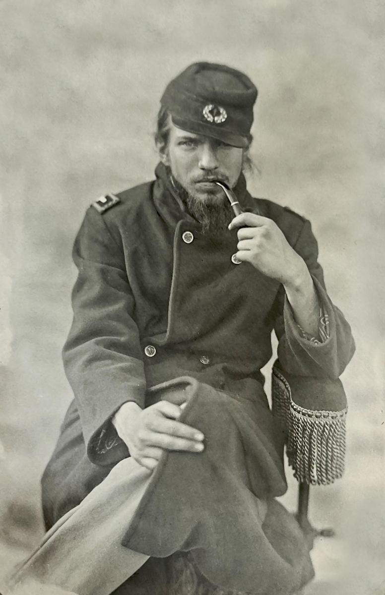 A man in a military uniform with a beard and long hair sits holding a pipe in his mouth.