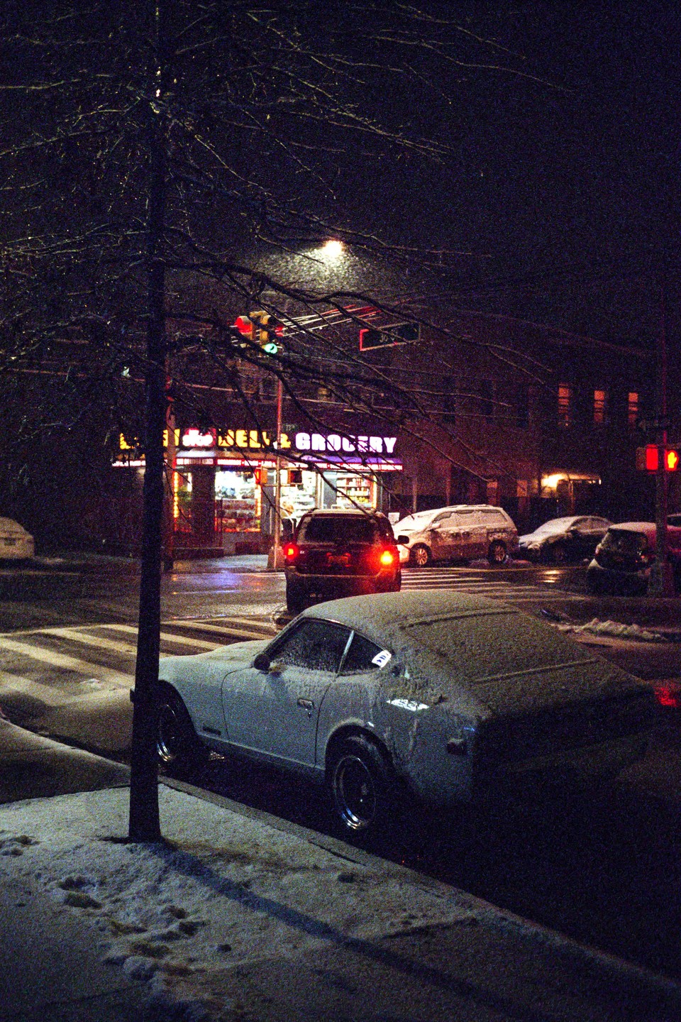 A snow-covered car is parked on a street at night, with a brightly lit grocery store in the background.