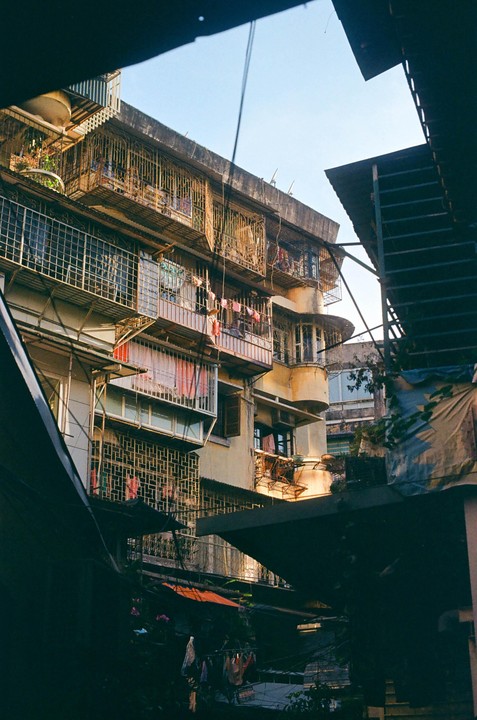 A tall, weathered building with many balconies and clotheslines is viewed from below, framed by dark structures.