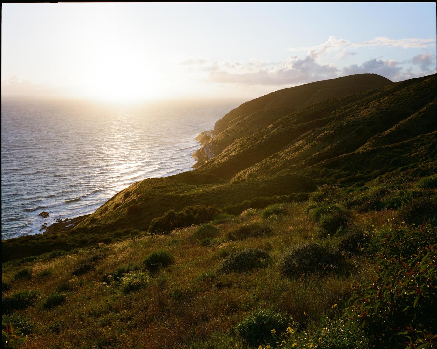 The sun sets over the Pacific Ocean, illuminating the water and a winding road along the PCH coastline.