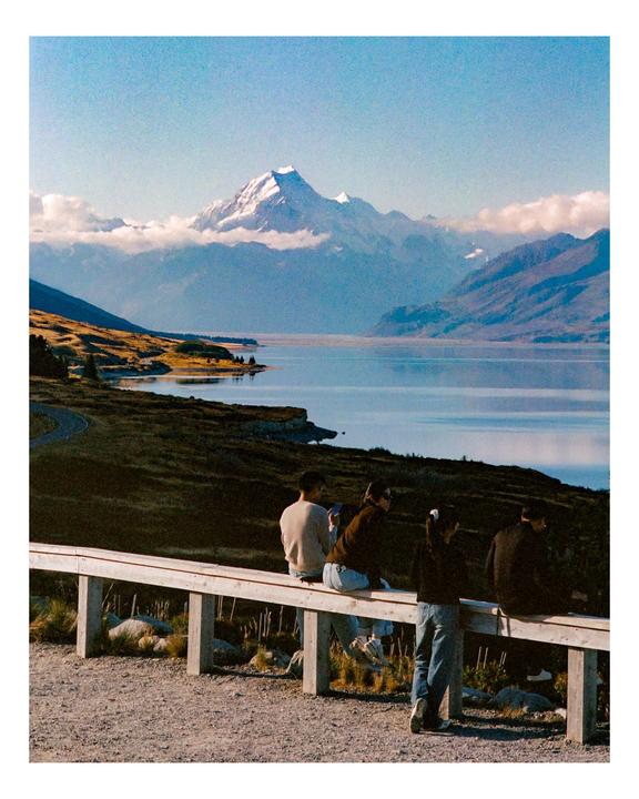 Four people sit on a wooden fence overlooking a lake and snow-capped mountains.