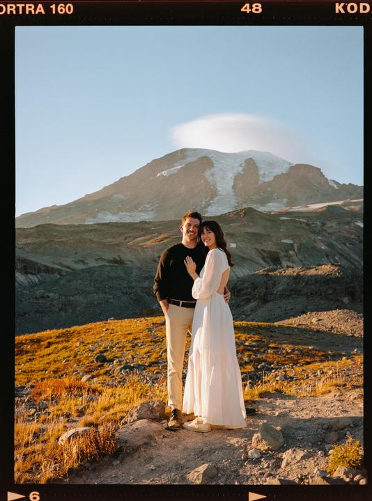 A couple stands on a rocky, grassy hill with a snow-capped mountain in the background.