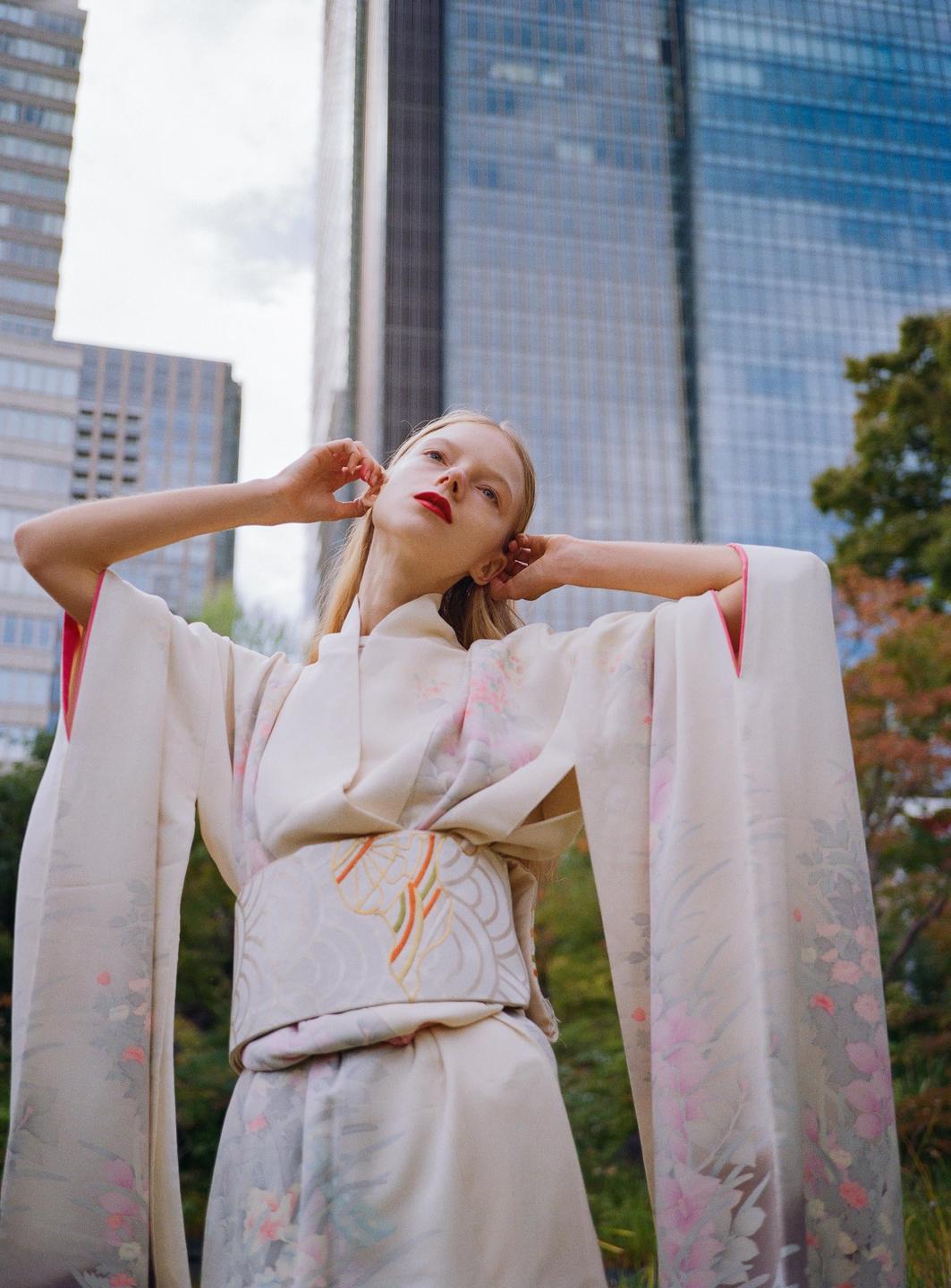 A woman in a kimono stands outdoors with tall buildings in the background.