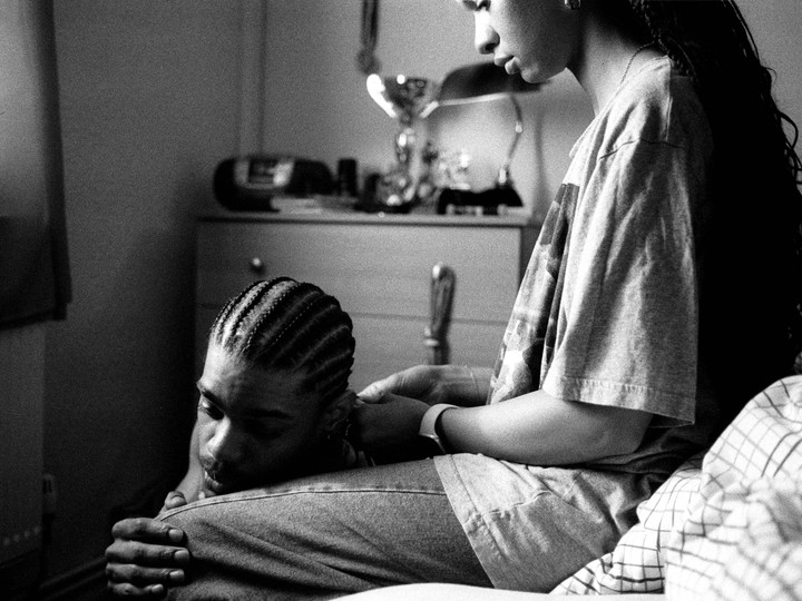 A woman sits on a bed with a man resting his head on her lap, while she touches his shoulder.