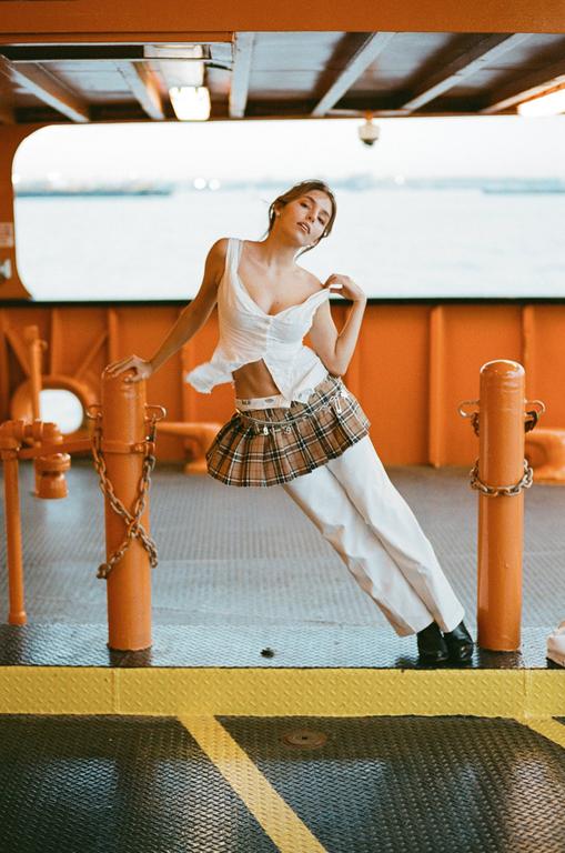 A woman in a white top and plaid skirt leans against an orange pole on a boat.