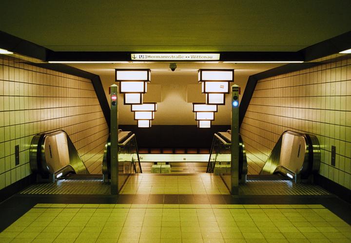 Two escalators descend into a subway station with tiled walls and illuminated signs.