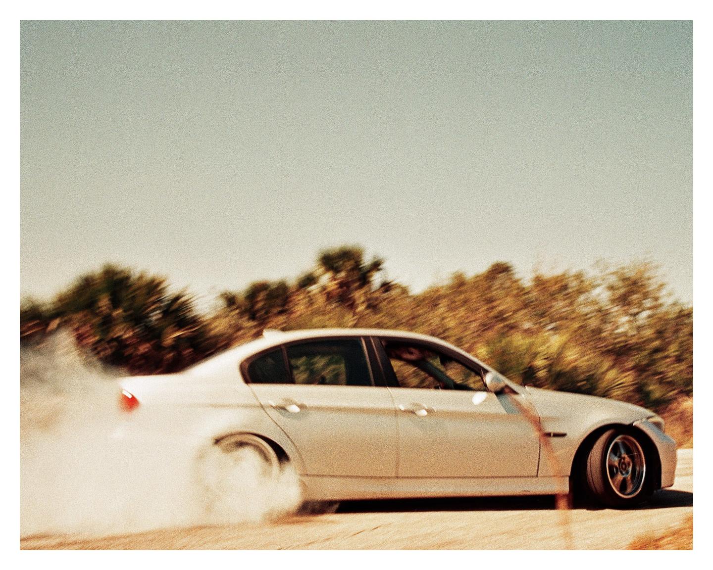 A white car drifts on a road, creating a cloud of smoke from its tires, with blurred trees in the background.