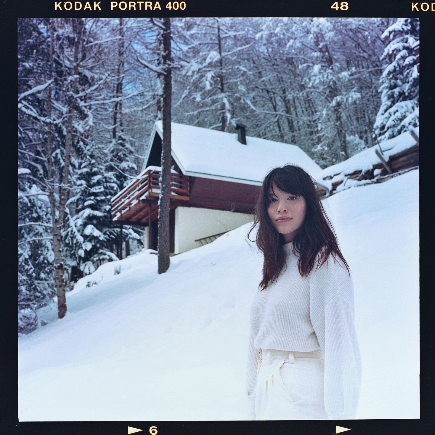 A woman stands in the snow in front of a chalet and snow-covered trees.