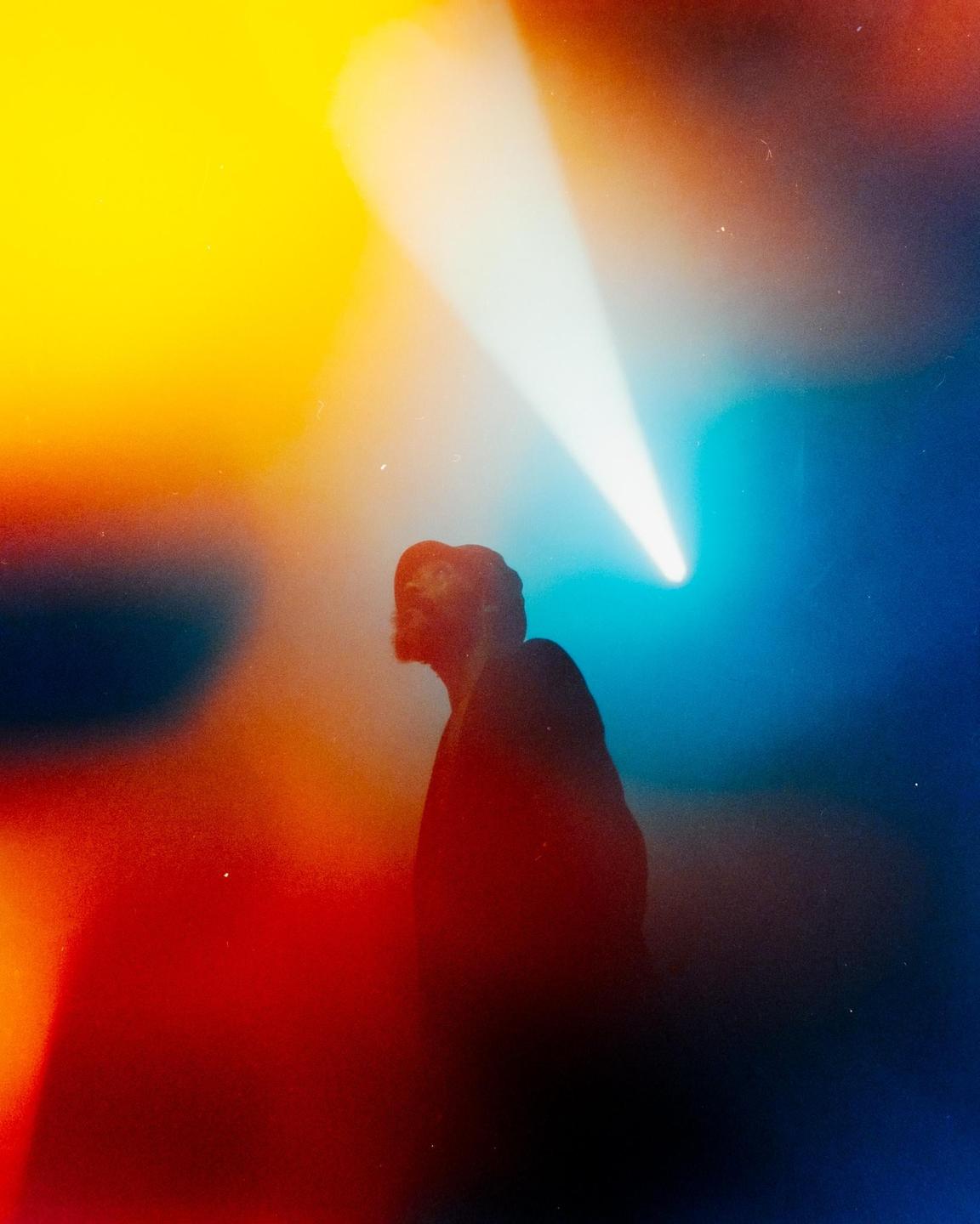 A man is illuminated by a spotlight in a colorful, hazy environment.
