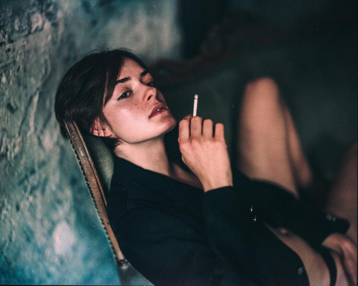 A woman with short hair is leaning back in a chair, holding a cigarette.