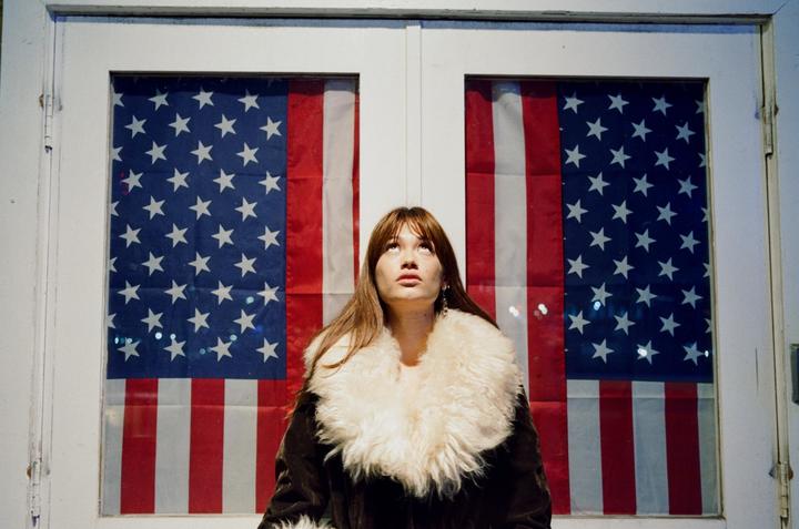 A woman in a fur-collared coat looks up, framed by two windows displaying American flags.