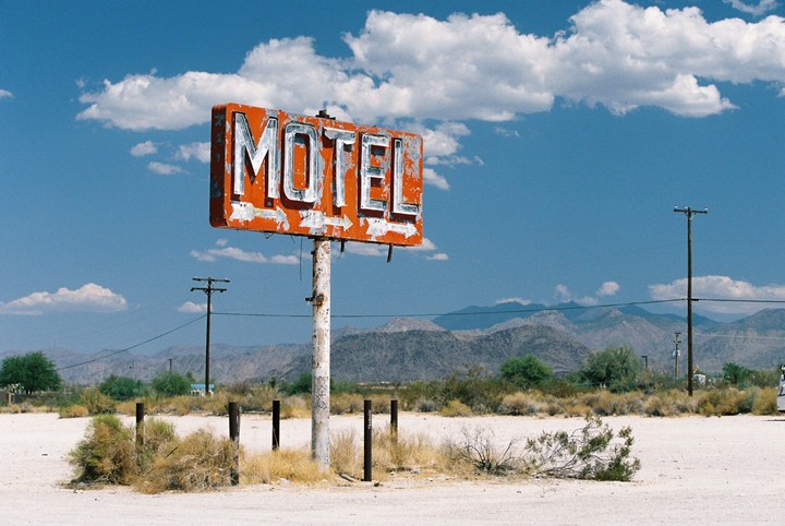 An old motel sign stands in a desert landscape with mountains in the background under a blue sky with clouds.