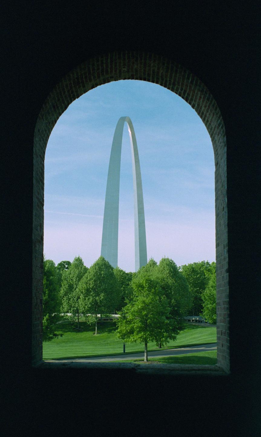 The Gateway Arch is visible through a dark brick archway, surrounded by green trees and grass under a clear blue sky.