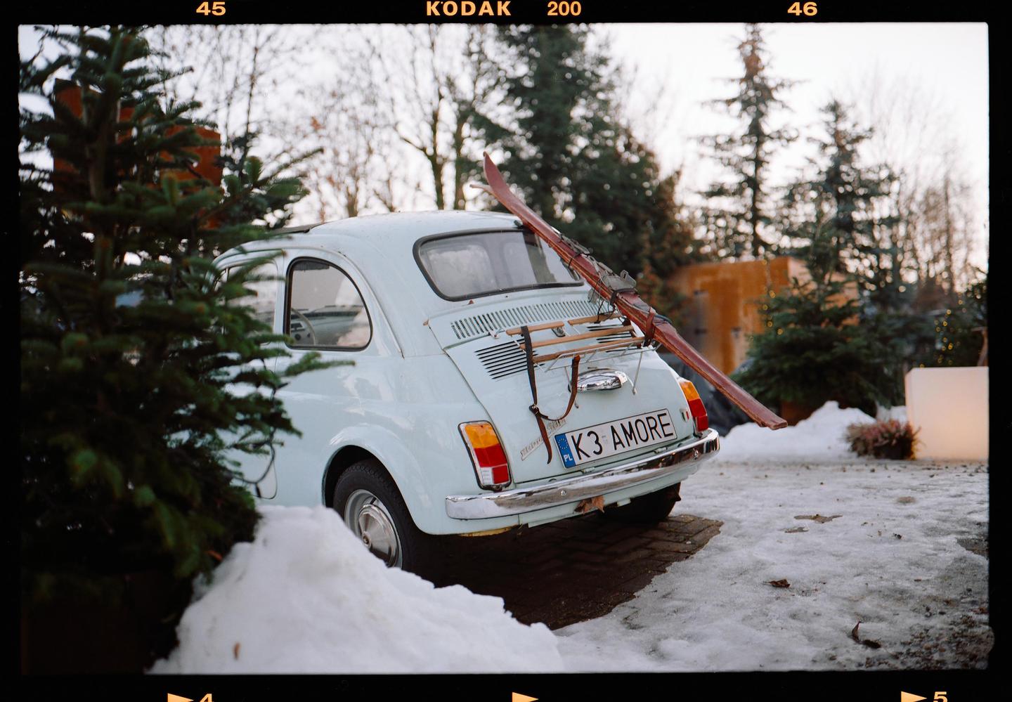 A light blue car with skis on its back is parked in a snowy area next to a pine tree.