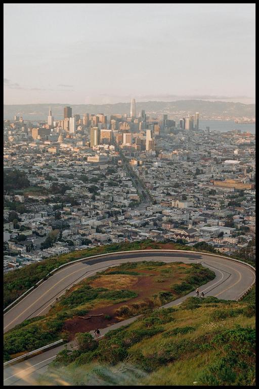A winding road leads up a hill overlooking a city skyline with a body of water in the distance.