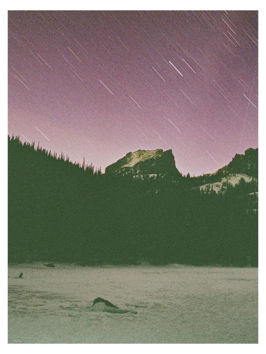 A long exposure captures star trails over a snow-covered landscape with mountains and a forest.
