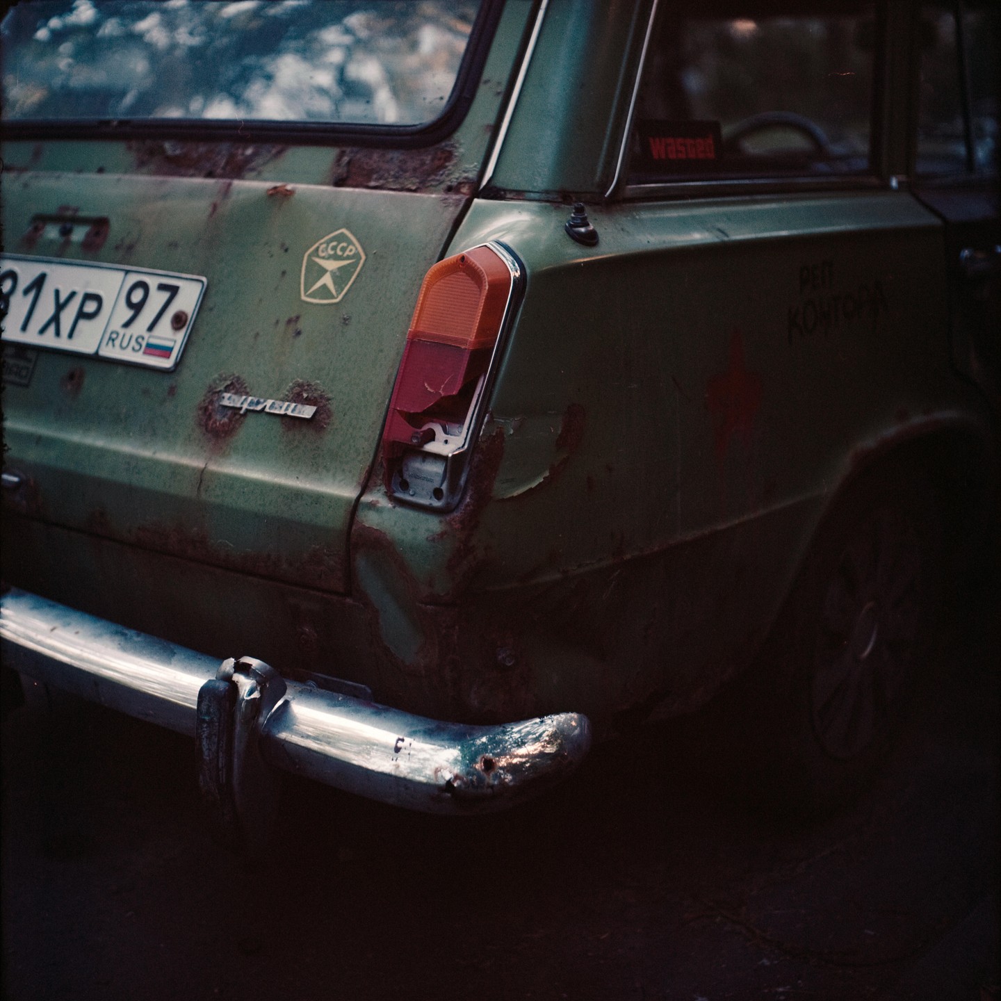 The rear of a rusty green station wagon with a broken taillight is parked in shadow.