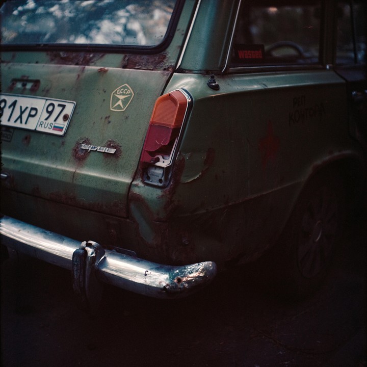 The rear of a rusty green station wagon with a broken taillight is parked in shadow.