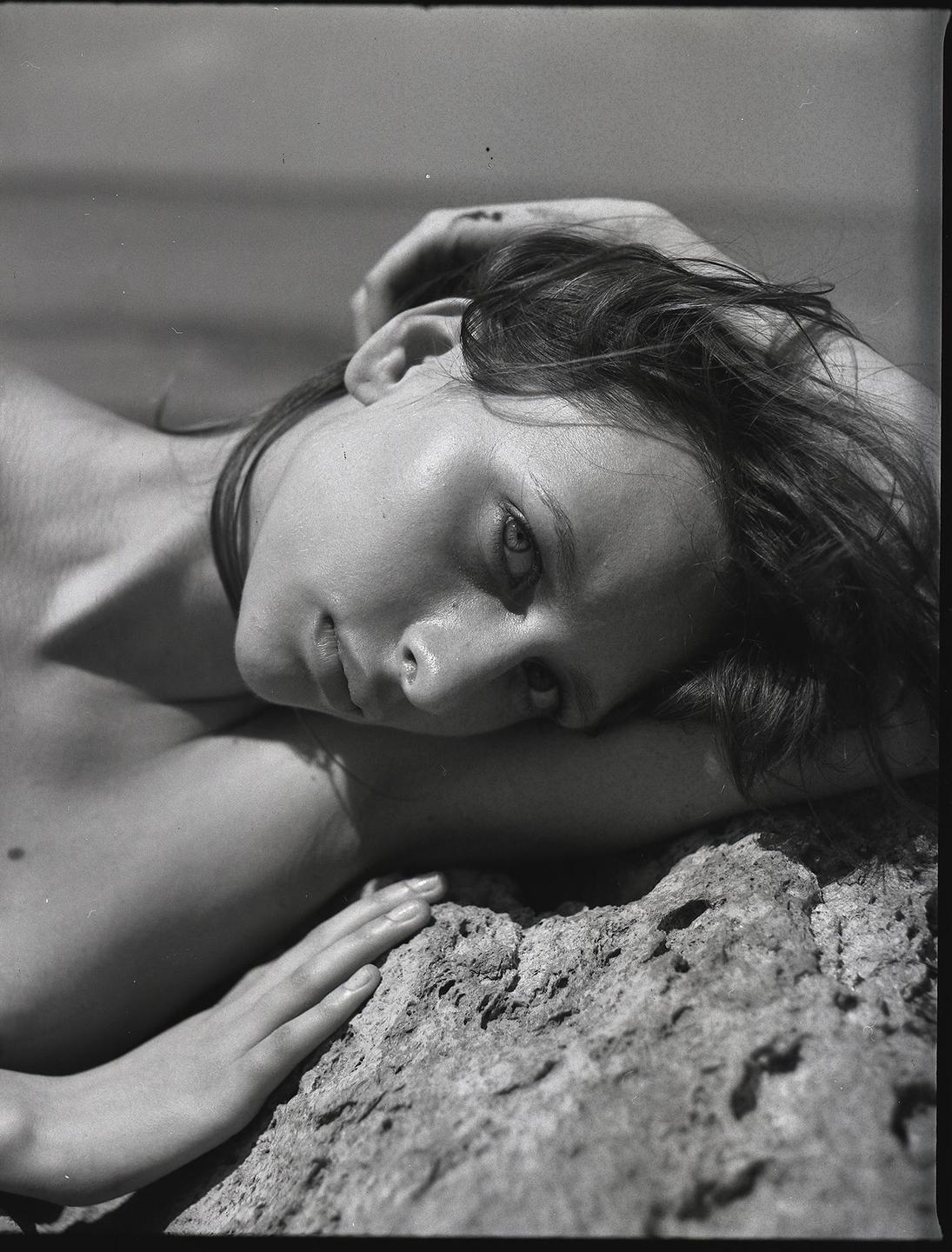A woman with dark hair is lying on a rock, looking towards the viewer.