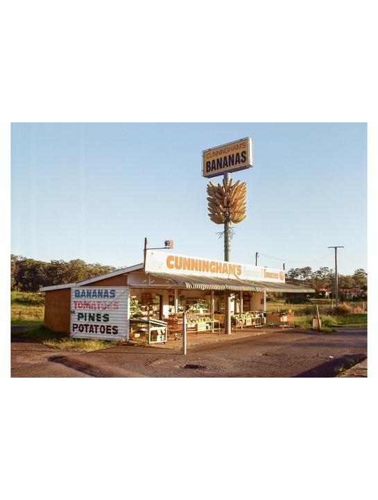 A roadside fruit stand with a large banana-shaped sign stands next to a dirt road.