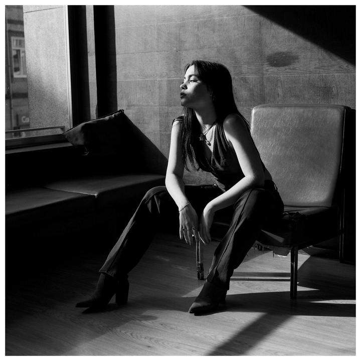 A woman sits on a chair next to a window, looking out with a thoughtful expression.