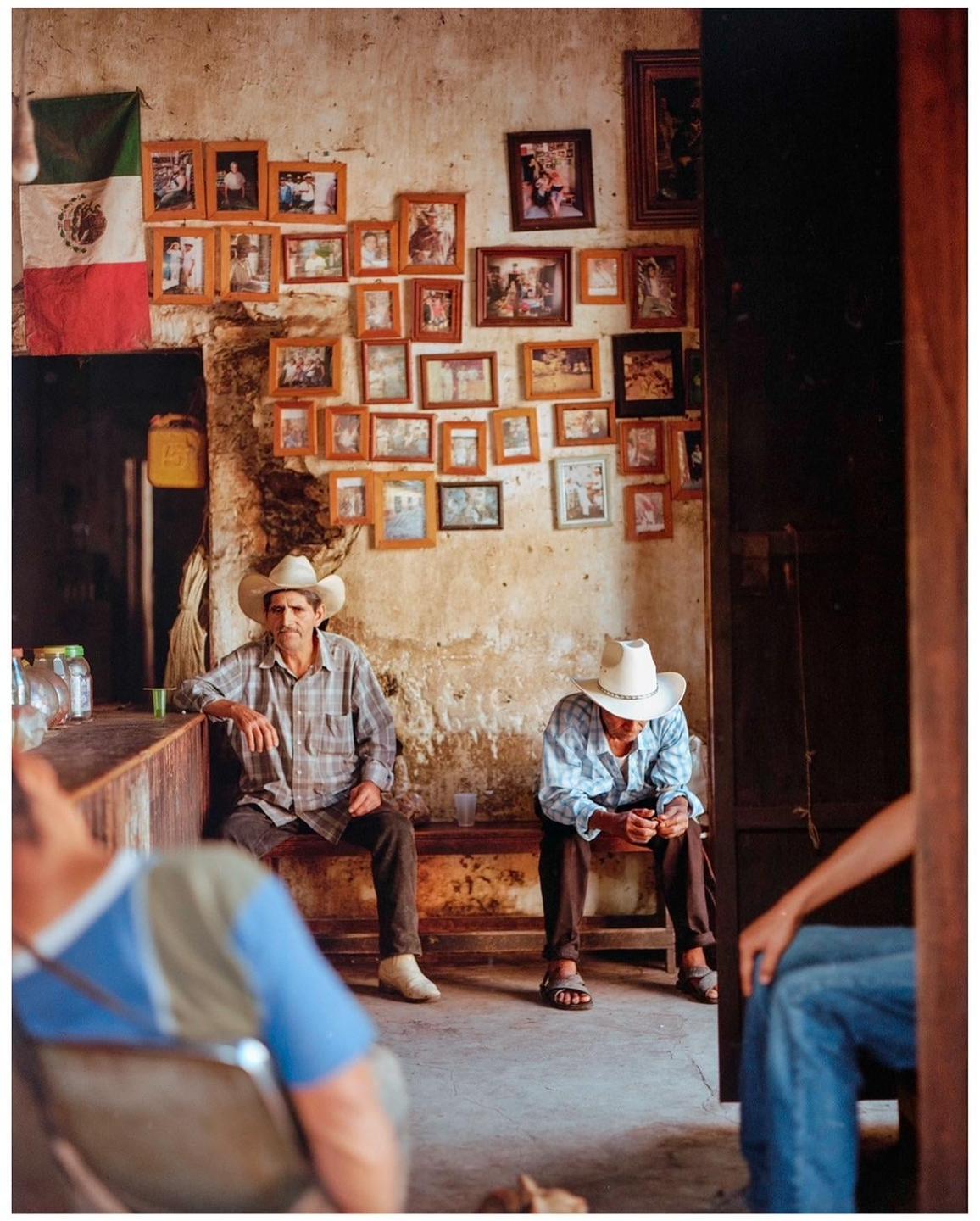 Two men wearing cowboy hats sit on a bench in a room with a Mexican flag and many framed photos on the wall.