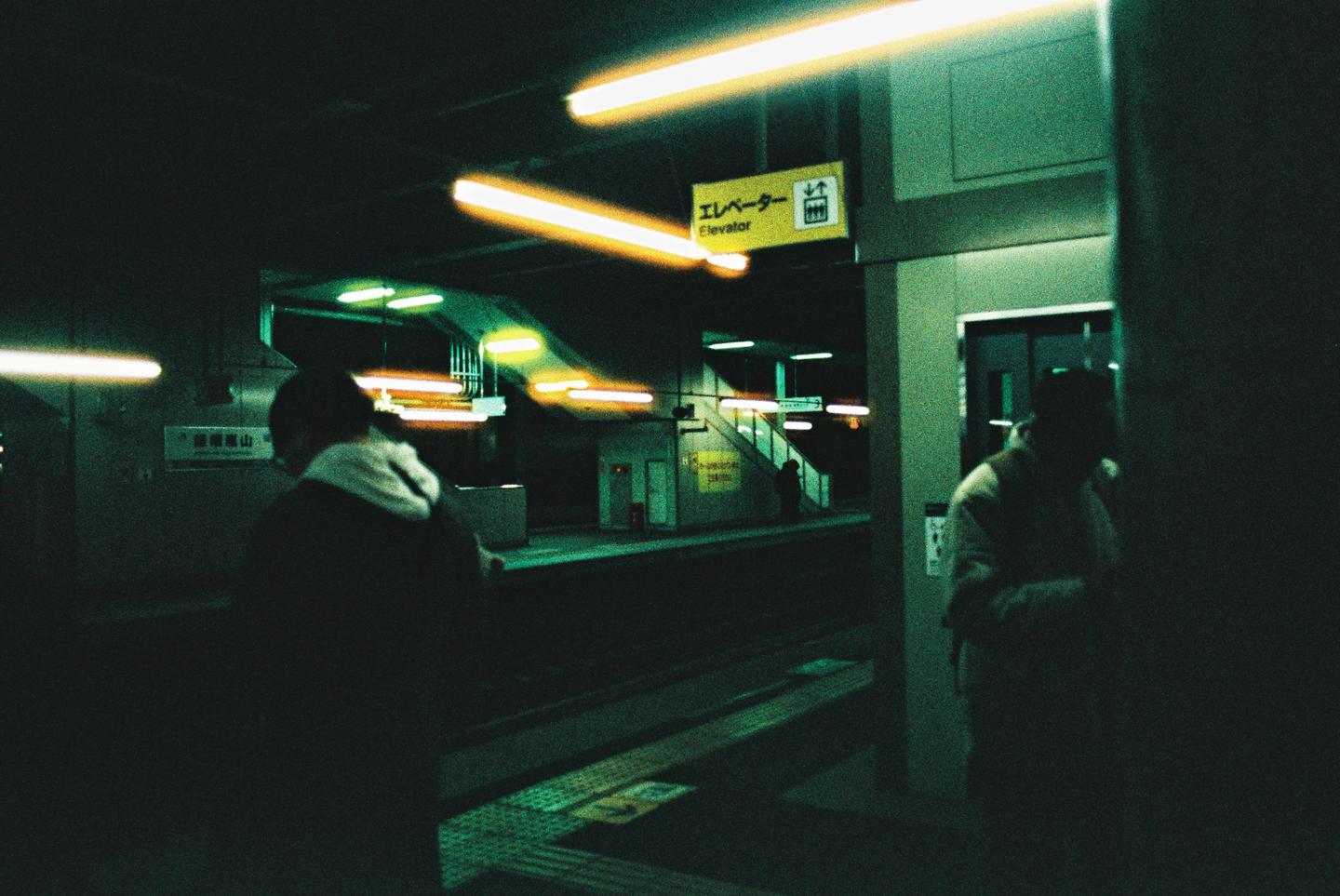 Two people stand on a dark subway platform with bright yellow and green lights.