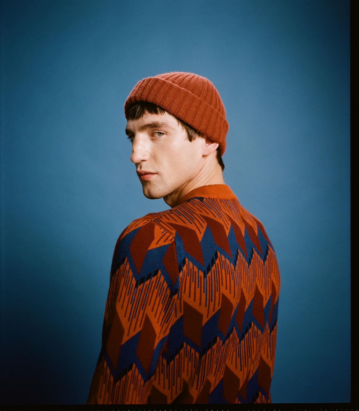A man in a patterned sweater and a beanie looks over his shoulder at the viewer.