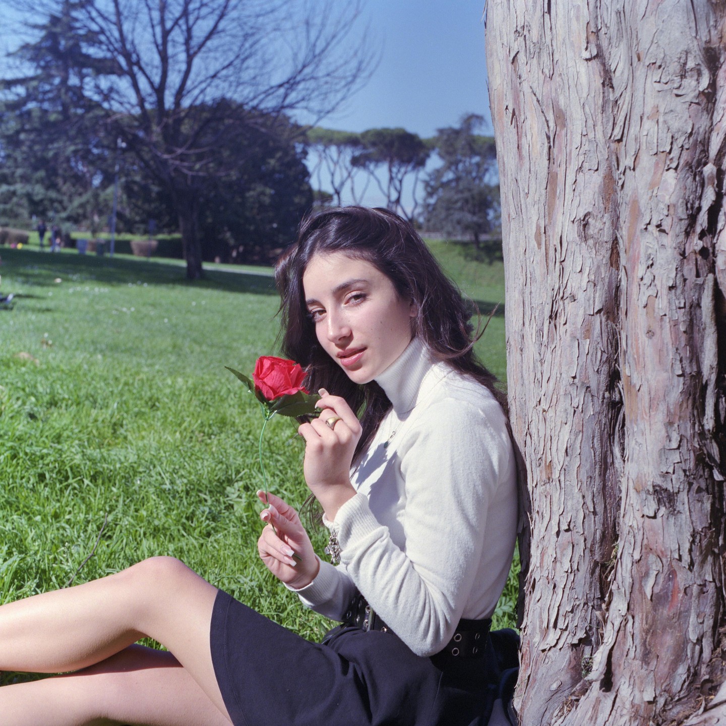 A woman sits on grass next to a tree trunk, holding a red rose.