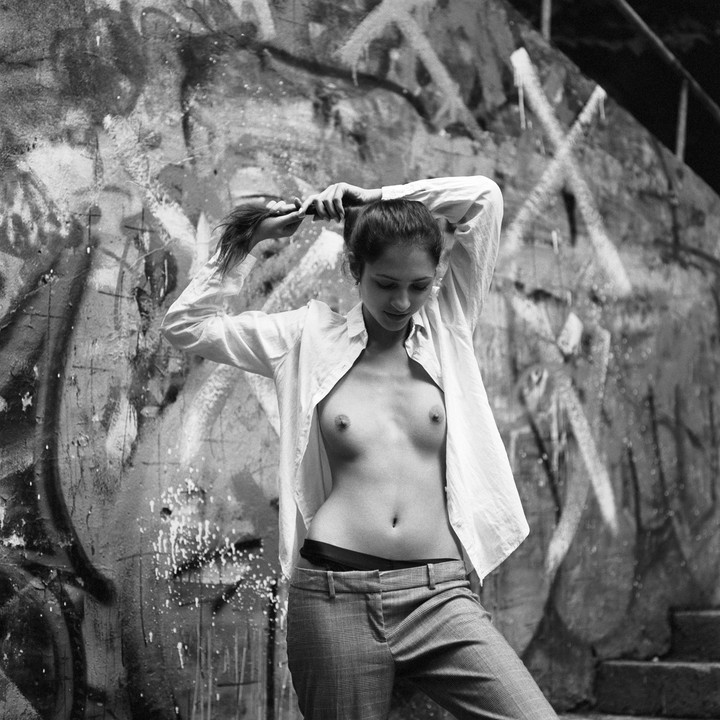 A woman with an open shirt and trousers stands in front of a graffiti-covered wall, pulling her hair up.